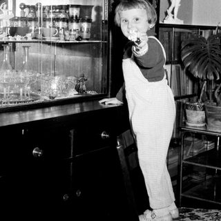 Boy in overalls stands on a chair near a cabinet.