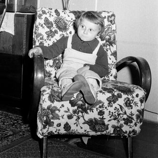 Here's a brief summary of the image: Young boy sits in a patterned chair, looking downwards.