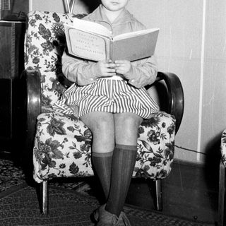 Here's a brief summary of the image: Girl sits reading a book in a vintage armchair.