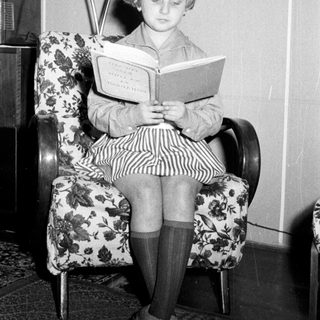 Here's a brief summary of the image: Girl sits reading a book in a vintage armchair.