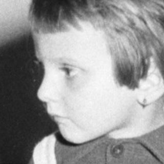 Here's a brief summary of the image: Child stands near a cake, wearing overalls.