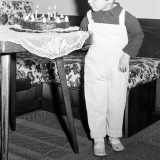 Here's a brief summary of the image: Child stands near a cake, wearing overalls.