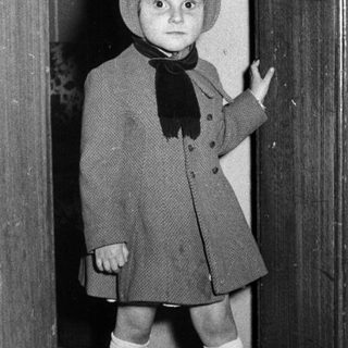 Here's a brief summary of the image: Young child in vintage clothing stands in a doorway.