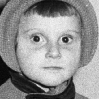 Here's a brief summary of the image: Young child in vintage clothing stands in a doorway.