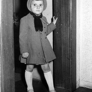 Here's a brief summary of the image: Young child in vintage clothing stands in a doorway.