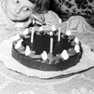 Child sits near a birthday cake with many candles.