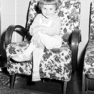 Here's a brief summary of the image: Child sits in a patterned armchair, smiling for the camera.