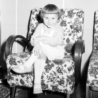 Here's a brief summary of the image: Child sits in a patterned armchair, smiling for the camera.