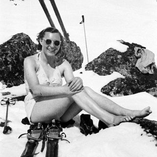 Woman in vintage swimsuit sits on snow with skis.