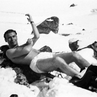 Man reclines in snow, wearing boots and shorts.