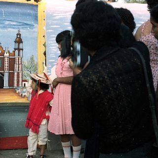 Girl in traditional dress stands before painted backdrop, being photographed.