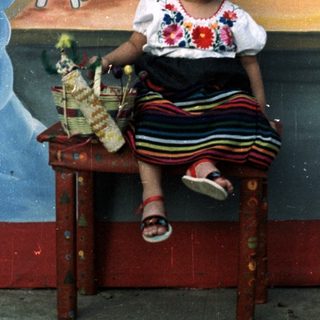 Here's a brief summary of the image: Girl in traditional dress sits before a painted church mural.