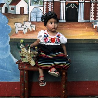 Here's a brief summary of the image: Girl in traditional dress sits before a painted church mural.