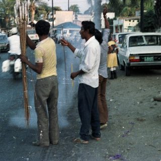 Men standing on road, burning sticks, onlookers watching.
