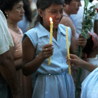 Girl holds candles, a child looks on in a crowd.