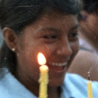 Girl holds candles, a child looks on in a crowd.