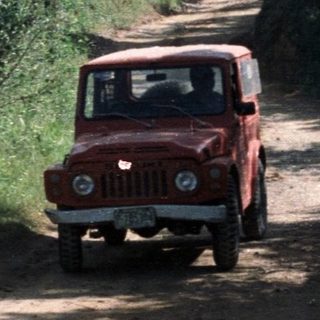 Red vehicle on dirt road, lush foliage, distant volcano.