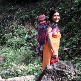 Woman with child stands near goats in a forested area.