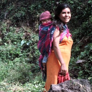 Woman with child stands near goats in a forested area.