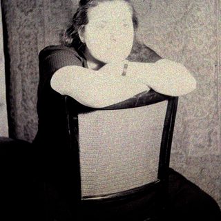 Here's a brief summary of the image: Woman seated in a chair, arms crossed, vintage photograph.