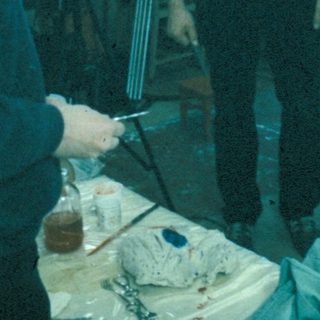 Man sculpts, two people observe in a workshop setting.