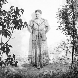 Woman stands outdoors near foliage and a wall.