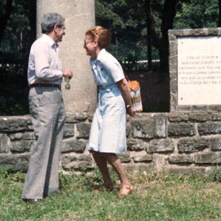 Couple stands near stone pillars and a plaque.