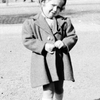 Here's a brief summary of the image: A young child stands outdoors in front of a building.