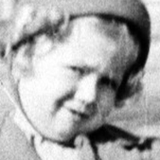 Here's a brief summary of the image: A young child stands outdoors in front of a building.