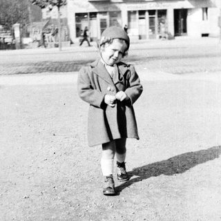 Here's a brief summary of the image: A young child stands outdoors in front of a building.