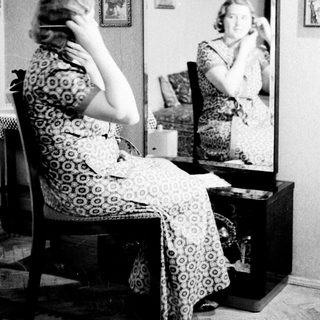 Woman sits before a mirror, seemingly applying makeup.