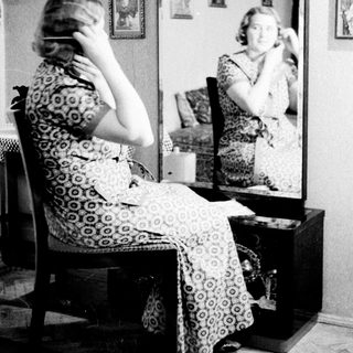 Woman sits before a mirror, seemingly applying makeup.