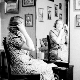 Woman sits before a mirror, seemingly applying makeup.