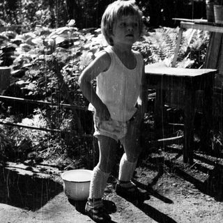Here's a brief summary of the image: Child stands in rain, appearing surprised in a garden.