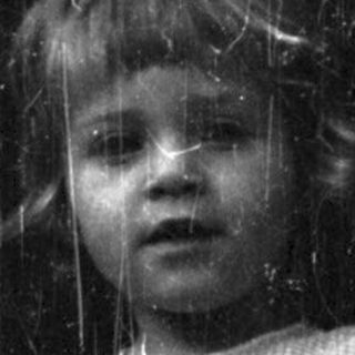 Here's a brief summary of the image: Girl stands near a door, peering out with a concerned expression.