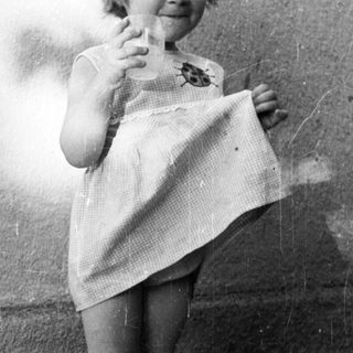 Here's a brief summary of the image: Young child stands outdoors, holding a dress, wearing shoes.