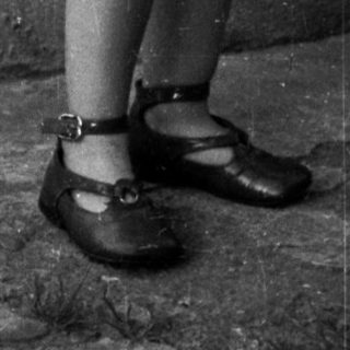Here's a brief summary of the image: Young child stands outdoors, holding a dress, wearing shoes.