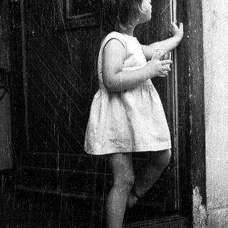 Here's a brief summary of the image: A young child stands reaching for a door handle.