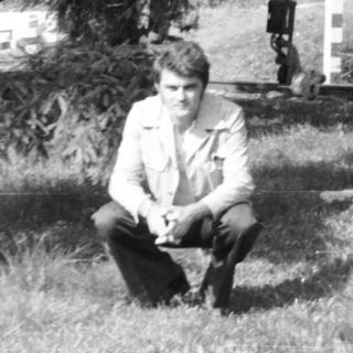 Man crouches in grassy area with trees and foliage.