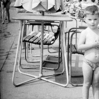 Boy stands near a pool with sunbathers in the background.