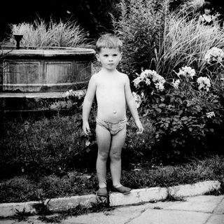 Here's a brief summary of the image: Boy stands outdoors, partially obscured by foliage and a figure.