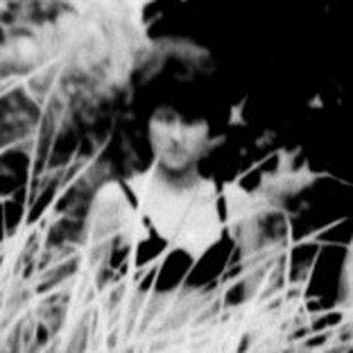 Here's a brief summary of the image: Boy stands outdoors, partially obscured by foliage and a figure.