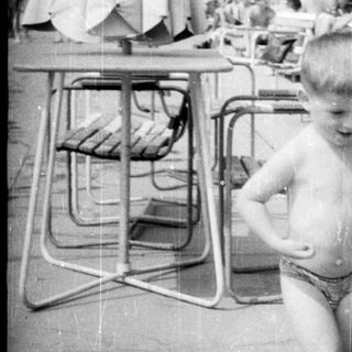 Boy stands near a pool with sunbathers in the background.