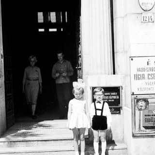 Two young children stand on steps; adults walk inside.