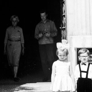 Two young children stand on steps; adults walk inside.