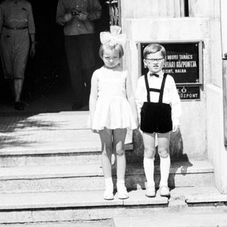 Two young children stand on steps; adults walk inside.
