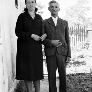 Older couple stands near a building in a rural setting.