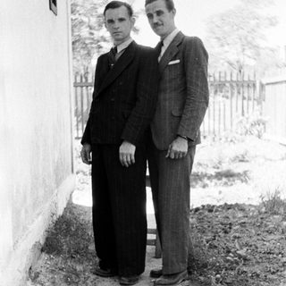 Two men in suits stand outdoors next to a wall.