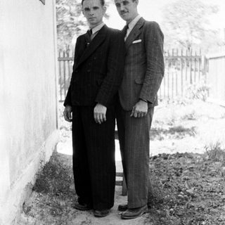 Two men in suits stand outdoors next to a wall.