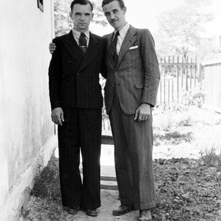 Two men in suits standing outdoors near a wall.